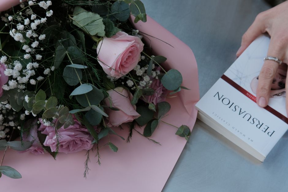 A floral arrangement featuring pale pink roses, complemented by sprigs of white baby's breath and eucalyptus leaves, all beautifully wrapped in soft pink paper. The bouquet is resting on a flat surfac