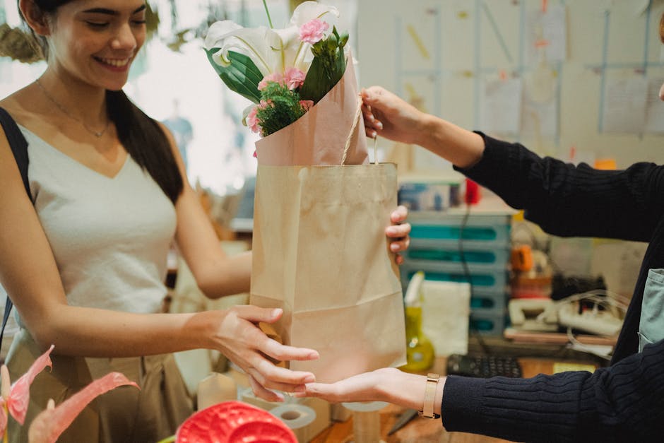 A woman with long dark hair, wearing a white sleeveless top, is smiling as she receives a floral gift from a florist at a flower shop. The florist, dressed in a dark sweater, hands over a brown paper