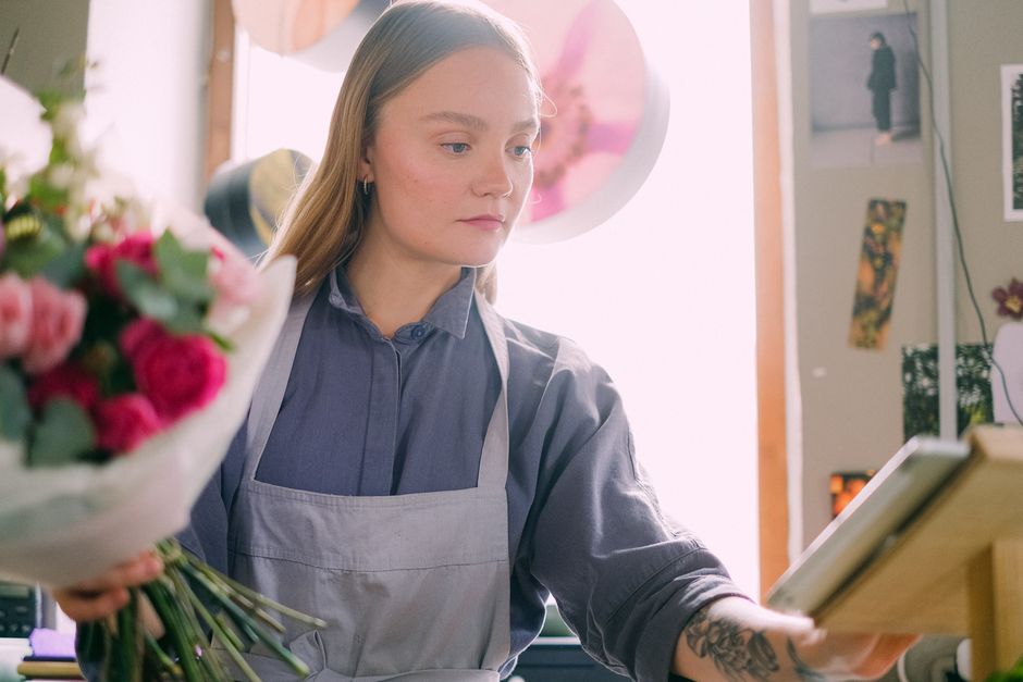 A young woman florist wearing a grey apron over a blue shirt is thoughtfully arranging a bouquet of fresh flowers inside a well-lit shop. She holds a bouquet of pink and white roses, complemented by g