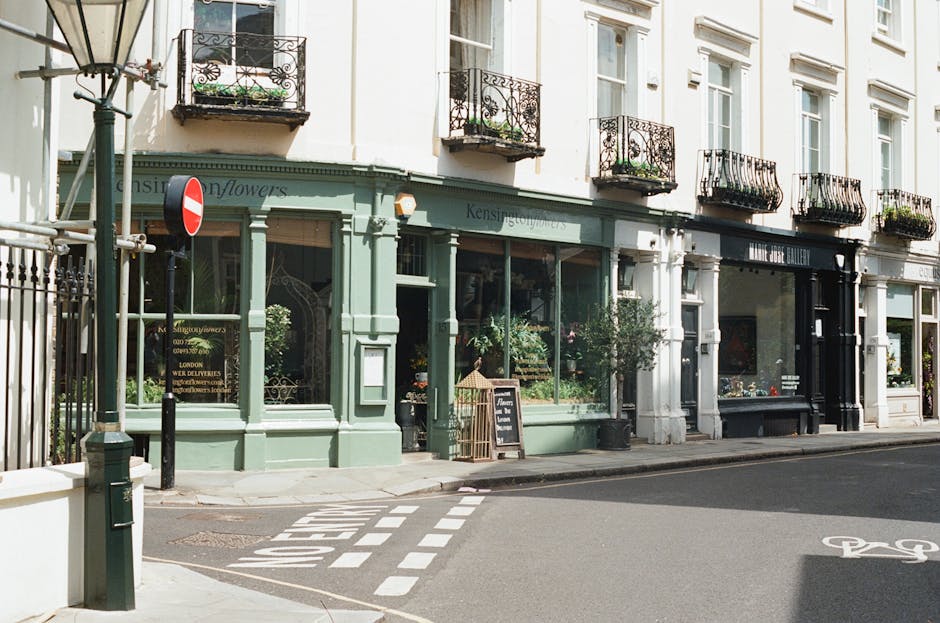 A row of charming florist shops with large display windows showcasing fresh floral arrangements, situated on a city street corner. The storefronts are painted in pastel hues, with one shop in light gr